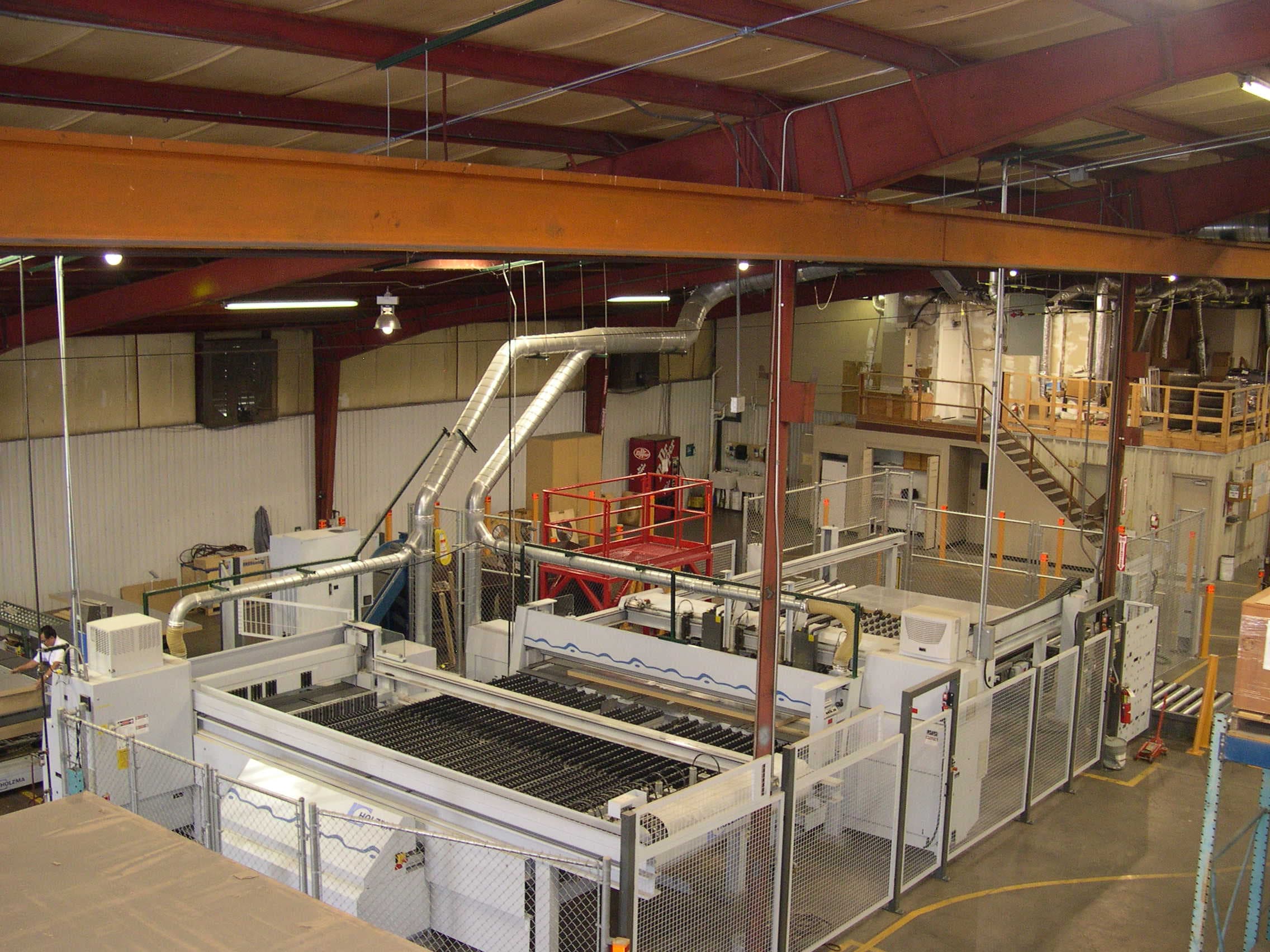 High-angle view of a manufacturing plant floor with large processing equipment, overhead ductwork, red structural steel, and industrial lighting.