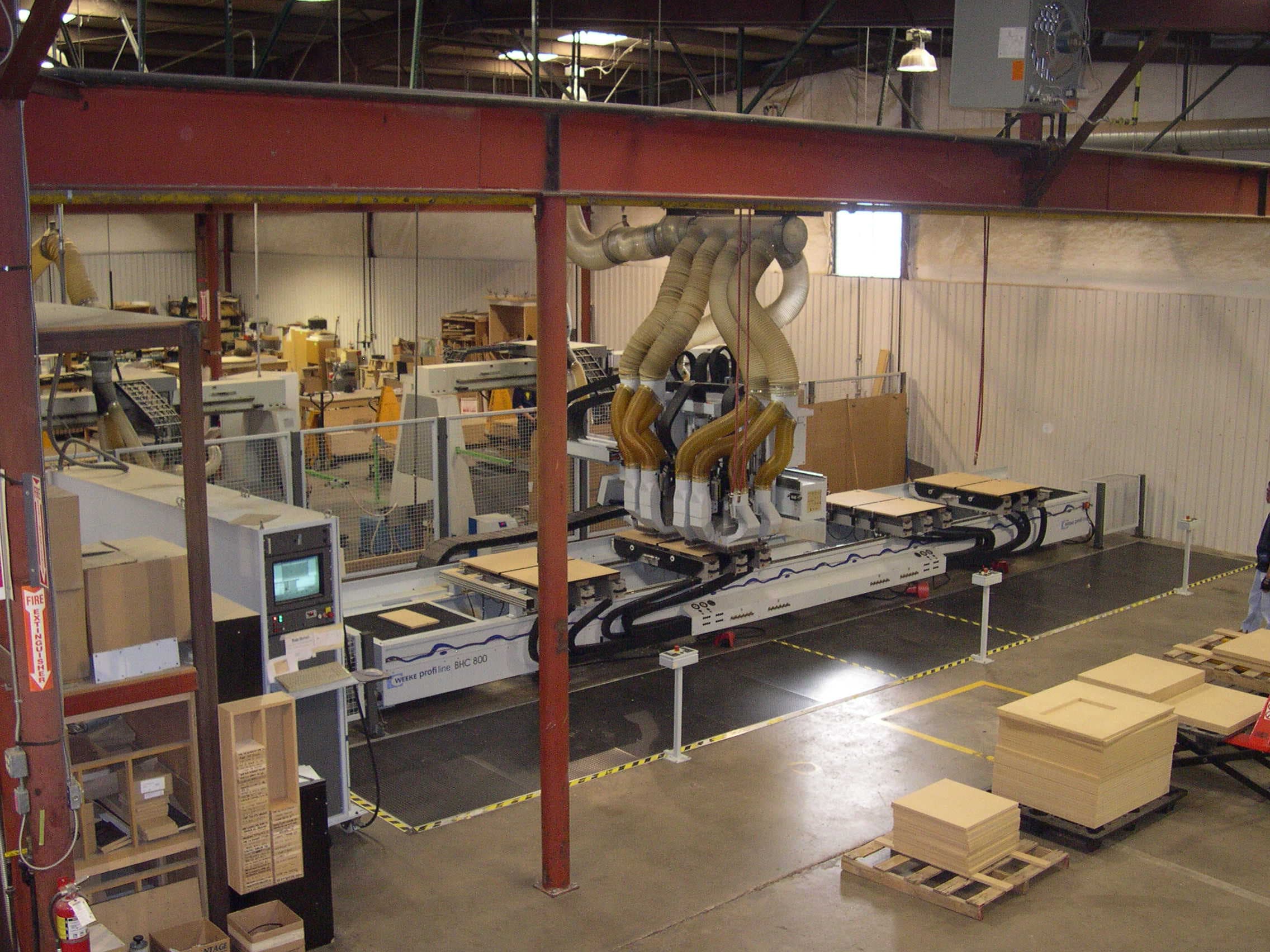 A large-scale automated CNC or panel machine with an overhead dust-collection system on a factory floor.