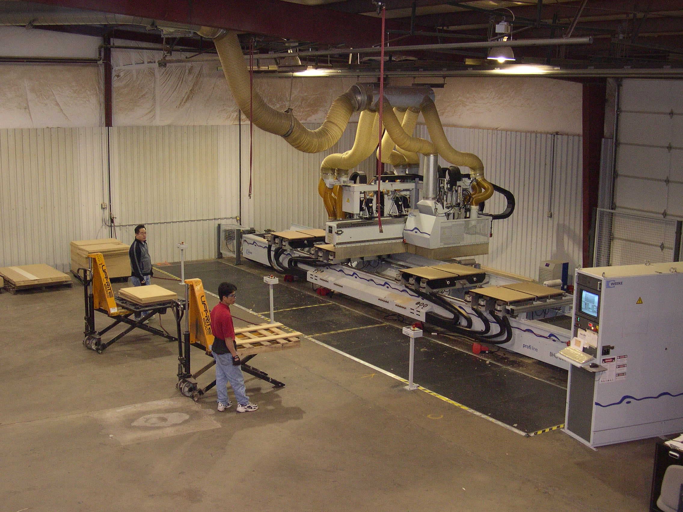 CNC woodworking center in operation, with a control console, vacuum hoses, and team members on the production floor.