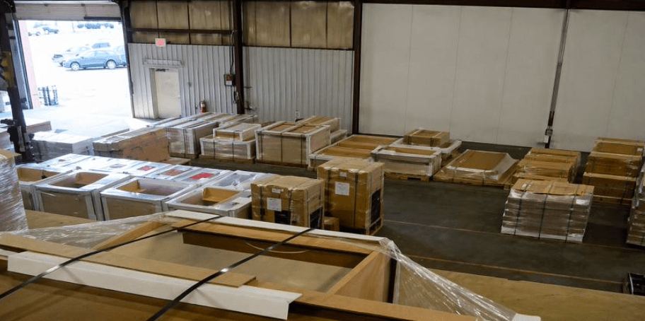Warehouse floor with rows of strapped, shrink-wrapped flat packs on pallets, loading bay door, and high-angle industrial view.