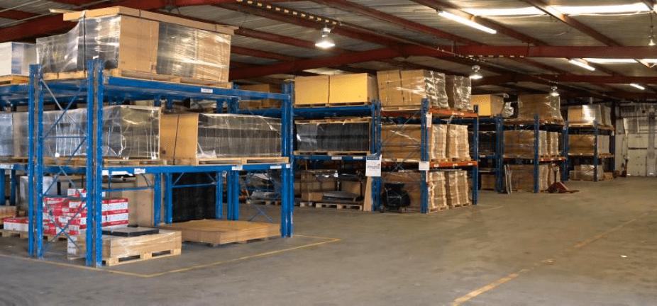 Organized distribution warehouse with blue pallet racks, stacked inventory, and yellow safety lines on a concrete floor.
