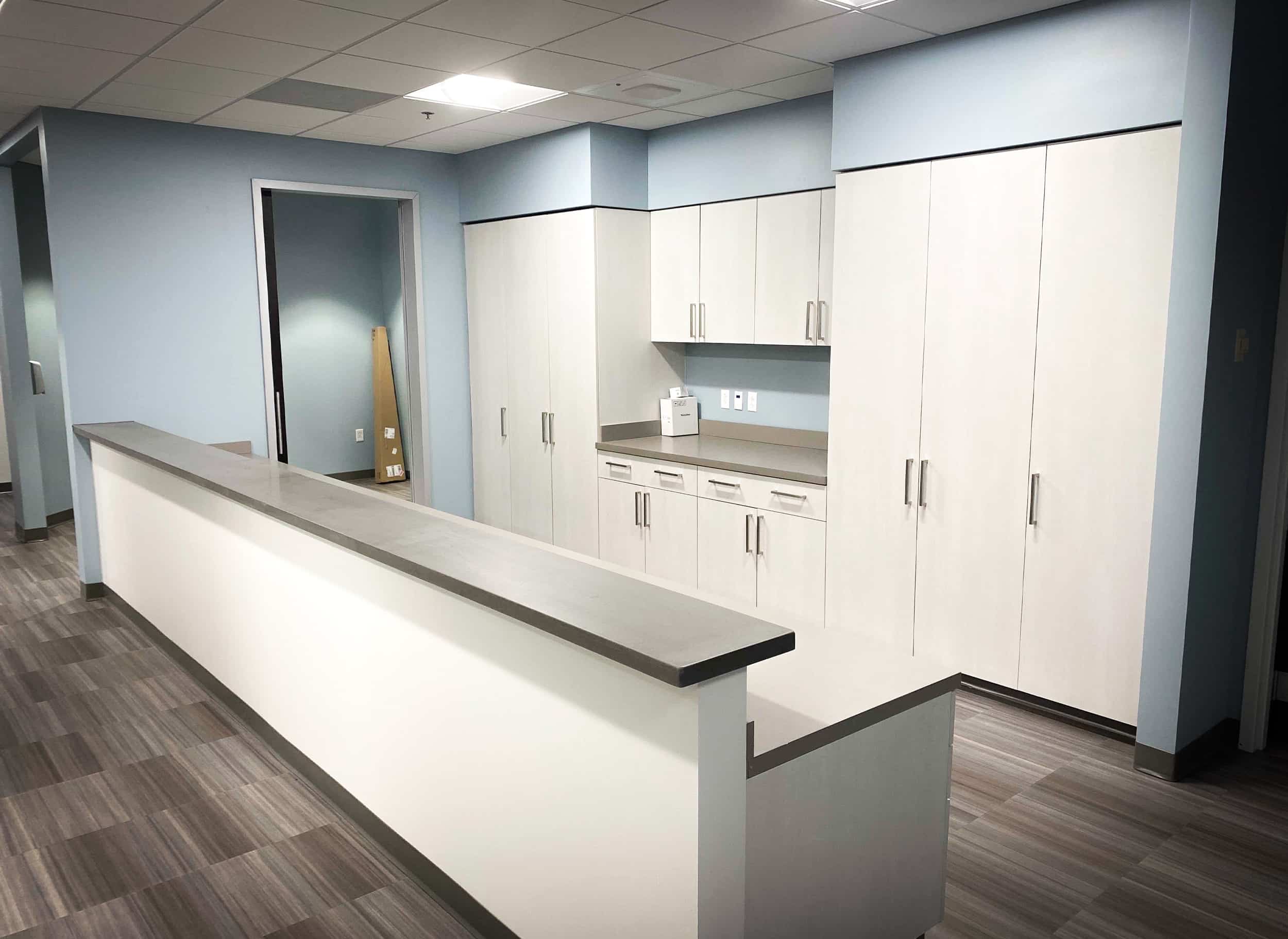 Healthcare nurse station with L-shaped counter, white casework, tall storage, and integrated workstation with grey countertops.